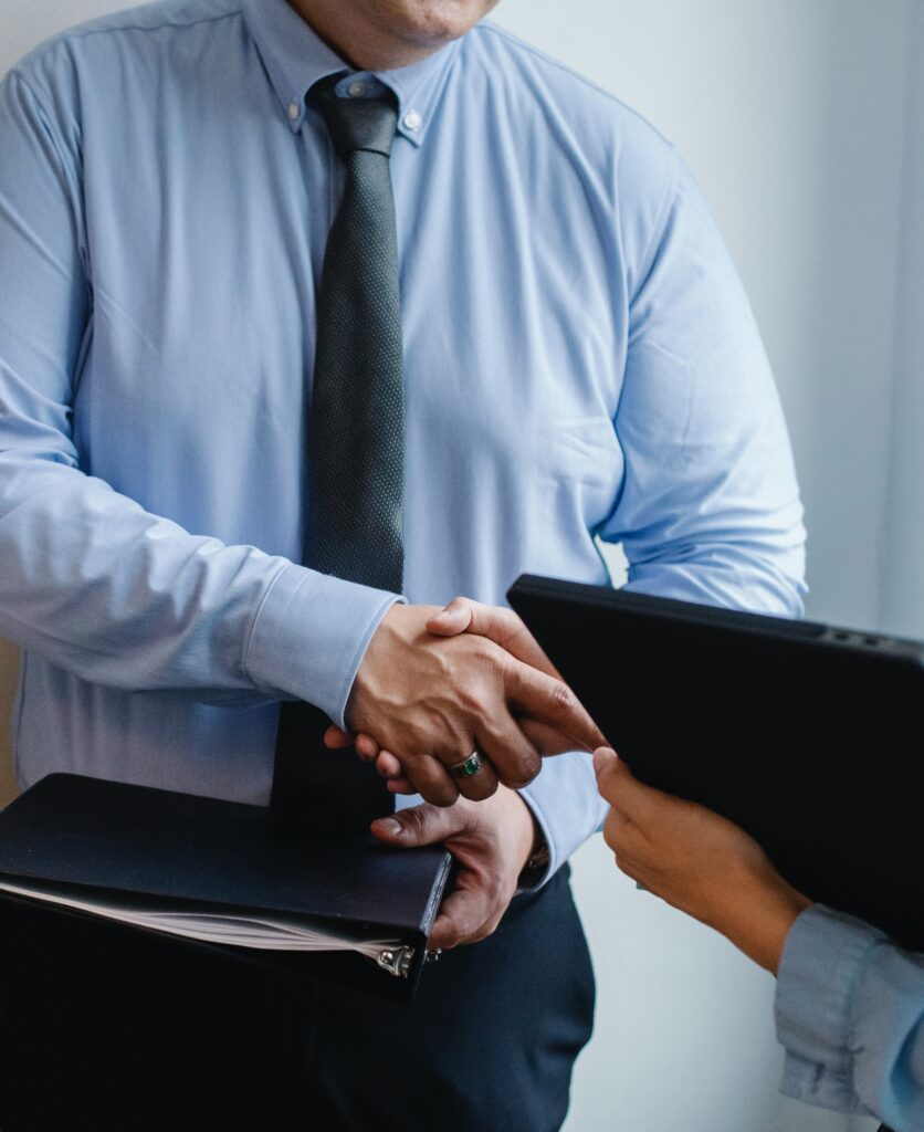 Un homme sert la main d'une personne dans le cadre d'une collaboration professionnelle. Il porte une chemise bleue et une cravate. Il a un classeur dans sa main.