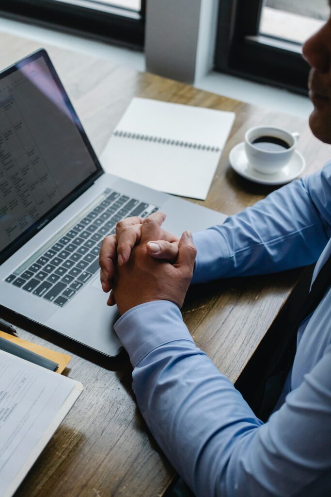 Un homme croise les mains sur son bureau. Il y a également un ordinateur portable et des dossiers professionnels. L'homme porte une chemise bleue.